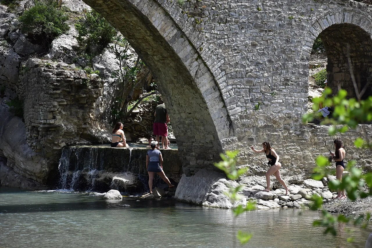 The old stone bridge at Bënjë near Përmet, where bathers gather by the water.