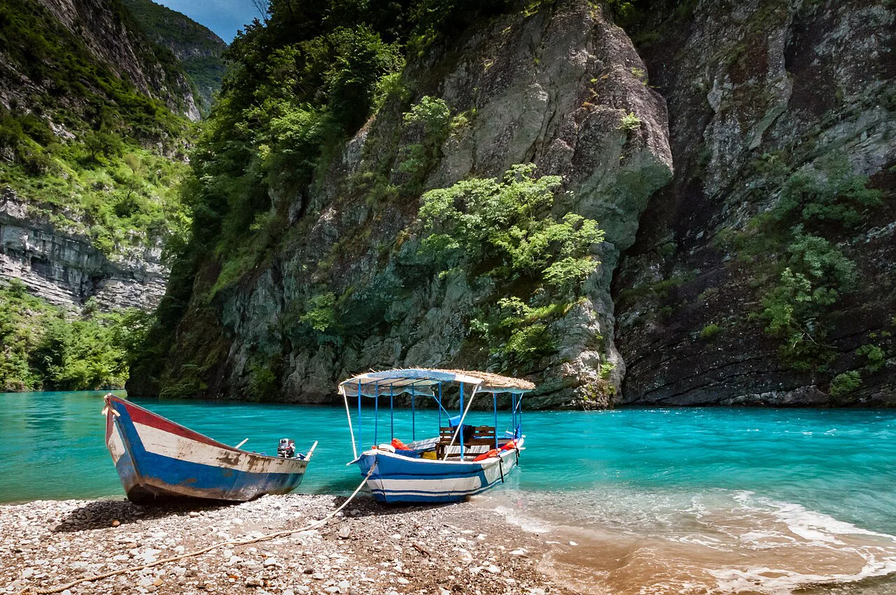 Boats moored on a pebble shore of Lake Koman beneath steep cliffs.
