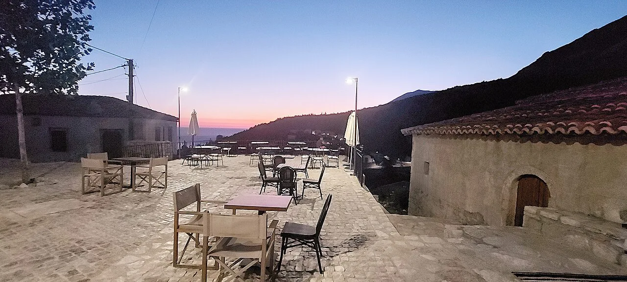 Café tables on the stone square in Dhërmi village at dusk.