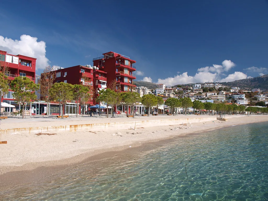 Clear water and the sandy town beach at Himarë, backed by seafront buildings.