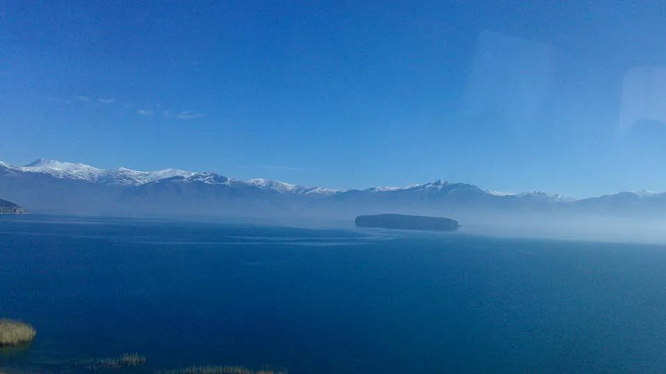 The calm blue water of Lake Prespa with snow-capped mountains beyond.