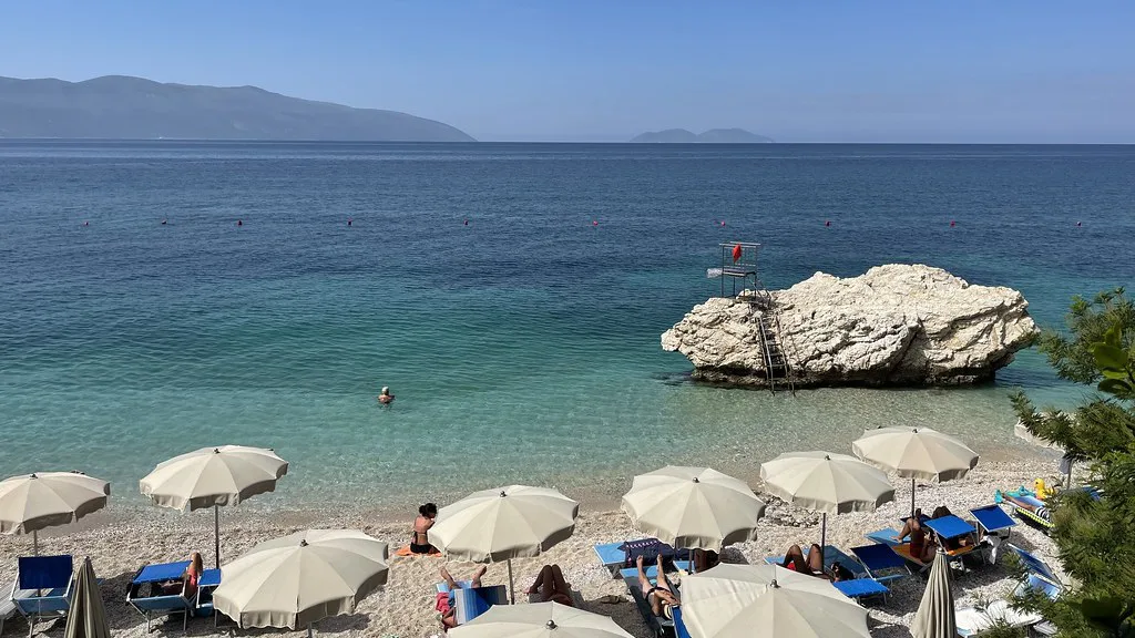 Parasols on a pebble beach near Vlorë with a large rock offshore and the Karaburun peninsula beyond.